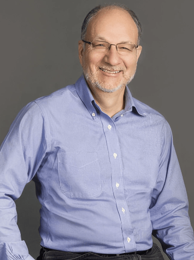 A smiling man with glasses wearing a light blue button-up shirt, seated against a gray background.