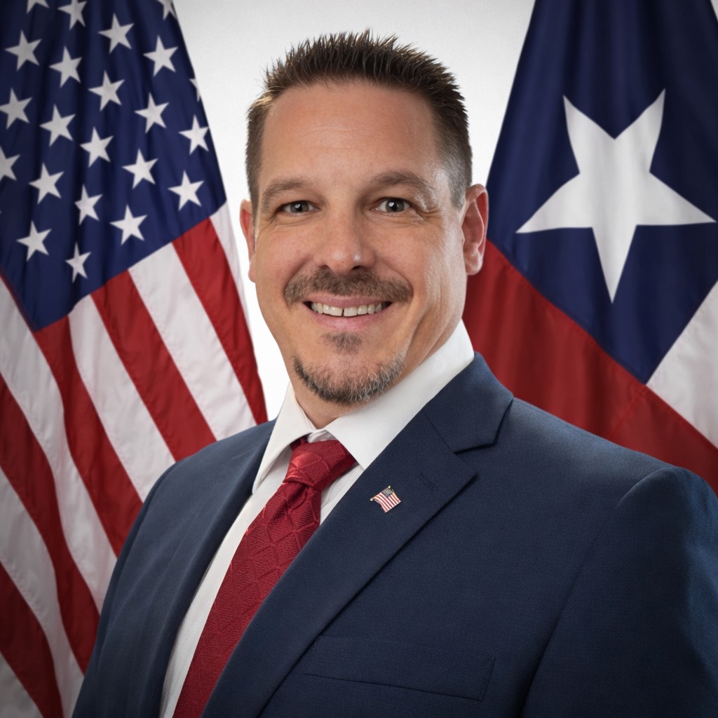Portrait of a smiling man in a suit with a red tie, standing in front of the American and Texas flags.