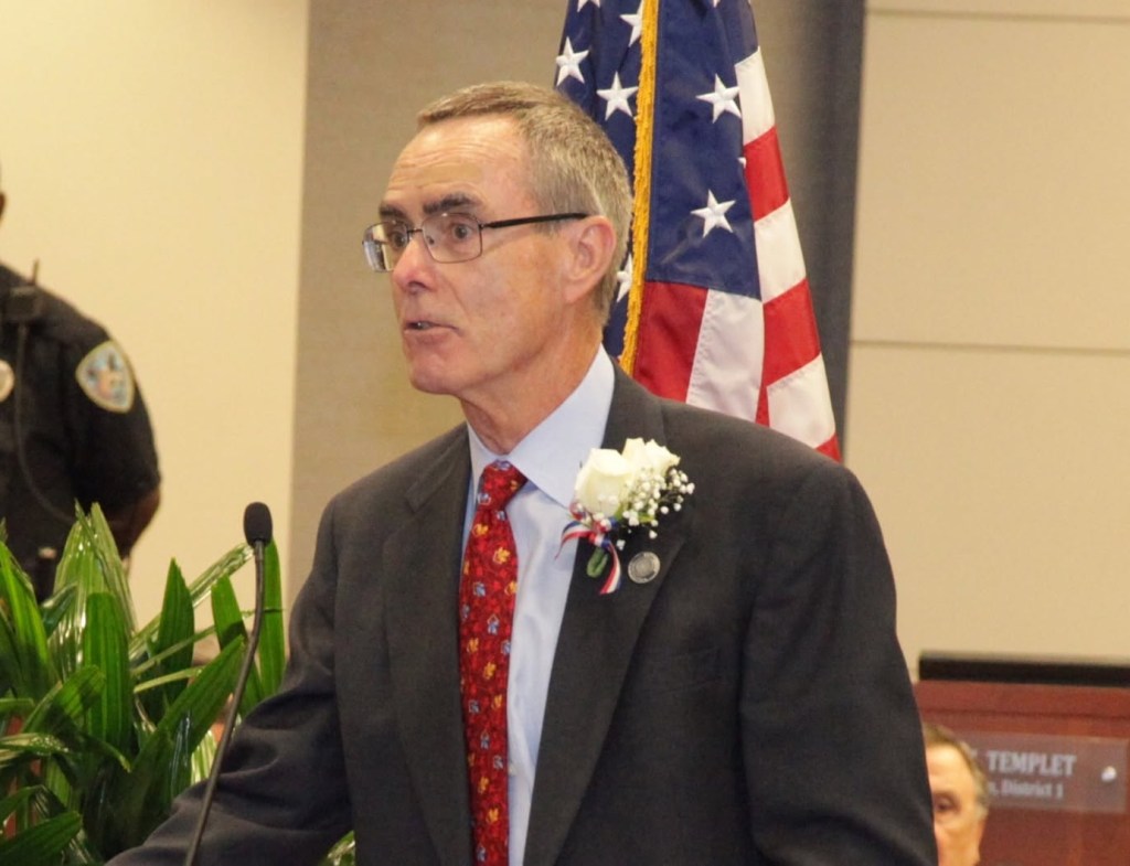 A man in a suit and glasses delivering a speech at a podium, with a bouquet on his lapel, and an American flag in the background.